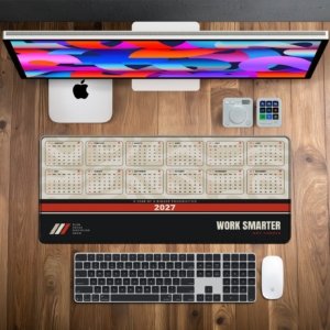 Work Smarter 2027 Calendar Desk Mat on a wooden desk with monitor, keyboard and mouse